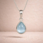Teardrop-shaped pendant with light blue natural aquamarine gemstone on a sterling silver chain against a soft pink background