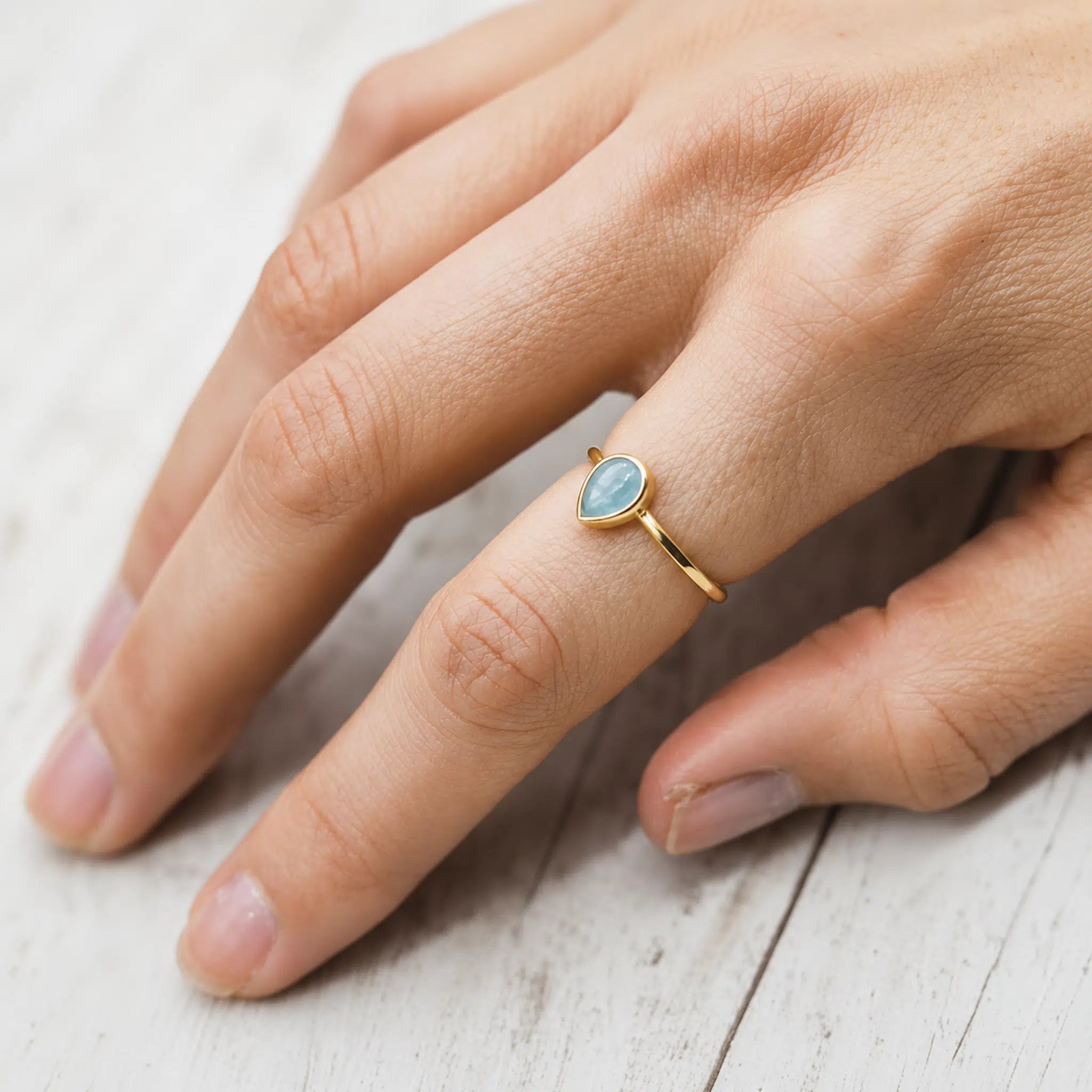 Hand wearing a gold march birthstone ring with a blue aquamarine gemstone on a light wooden surface