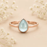Rose gold March Birthstone ring with a teardrop-shaped light blue natural aquamarine gemstone on a wooden surface.
