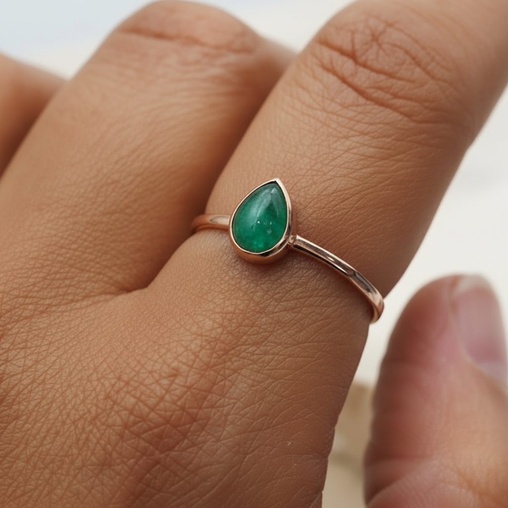 Rose Gold ring with a green tear drop shaped natural emerald gemstone on a person's finger against a neutral background