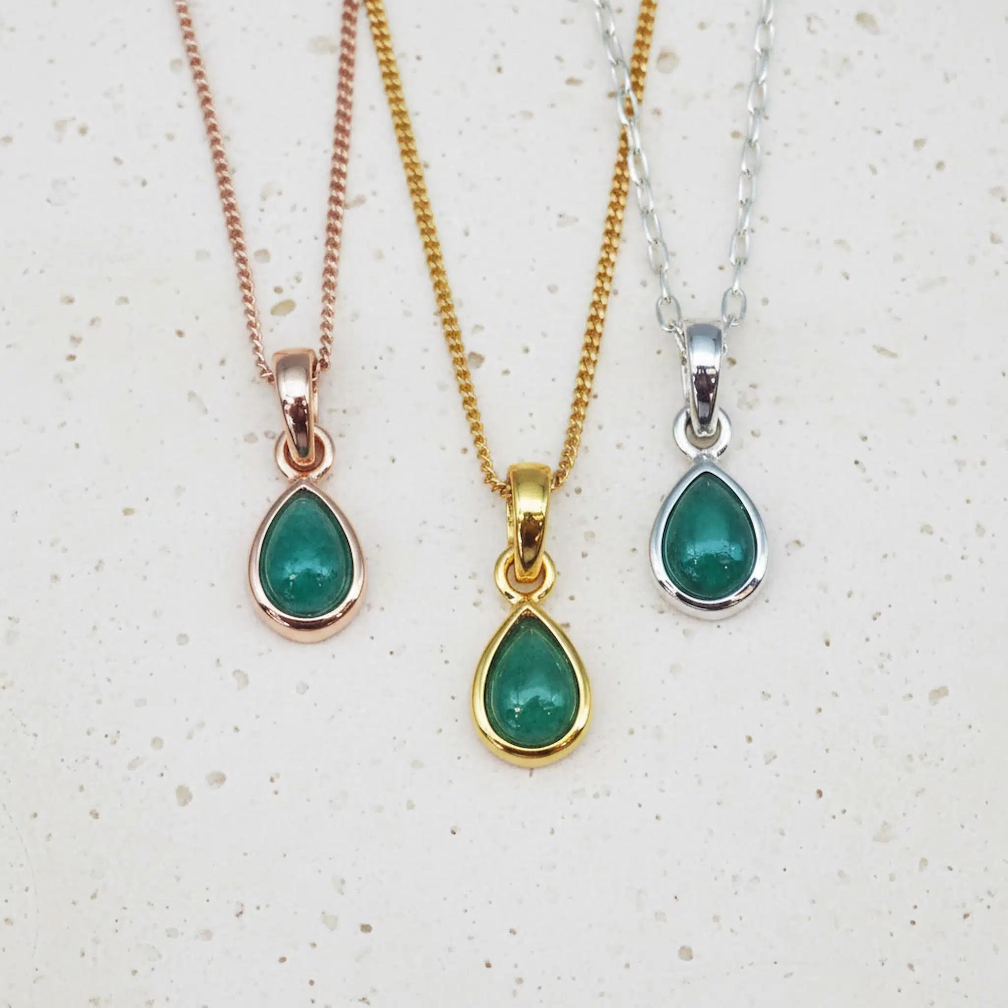Three may birthstone necklaces with teardrop-shaped natural emerald gemstone pendants on gold, rose gold, and silver chains against a light background.