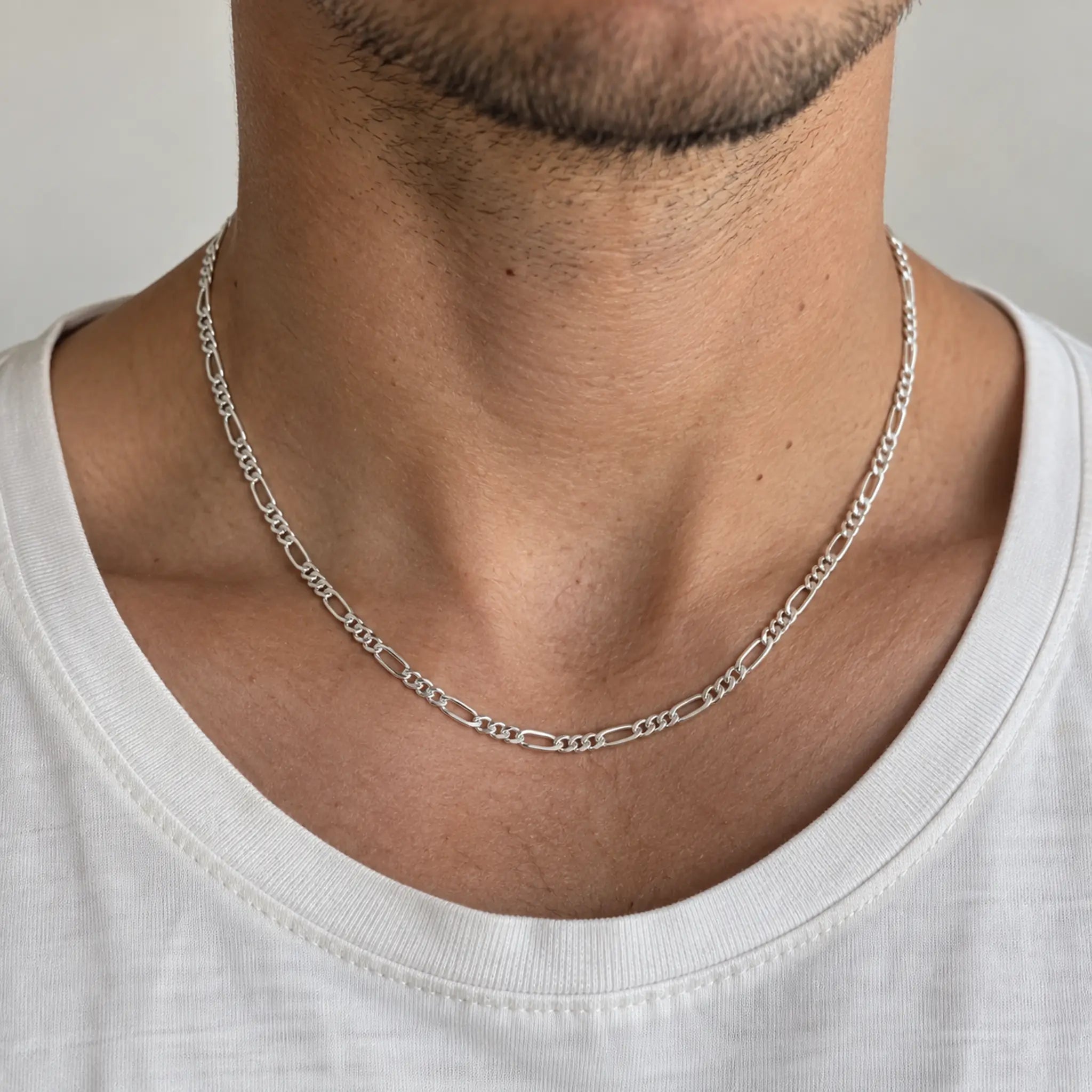 A close-up of a man's neck and chest wearing a silver Figaro chain necklace and a white crew-neck t-shirt.