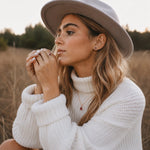 A woman wearing a light taupe wide-brim hat and a white ribbed knit turtleneck sweater, sitting in a dry grass field at sunset.