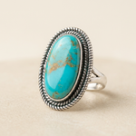 Mohave Turquoise Ring with a blue oval stone and gold veining in a rope-trimmed silver bezel, on a cream background