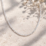 Delicate beaded natural rainbow moonstone necklace on a textured beige surface with small white flowers.