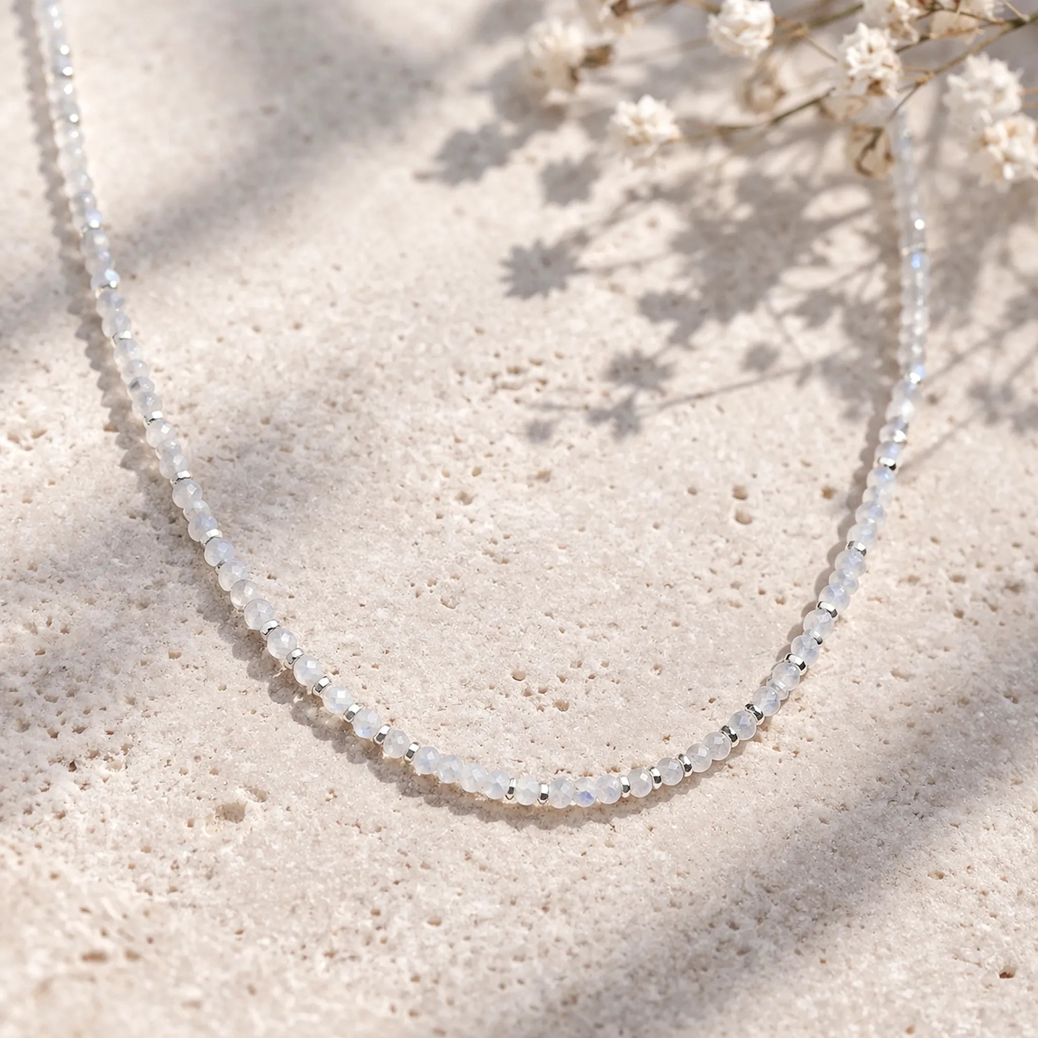 Delicate beaded natural rainbow moonstone necklace on a textured beige surface with small white flowers.