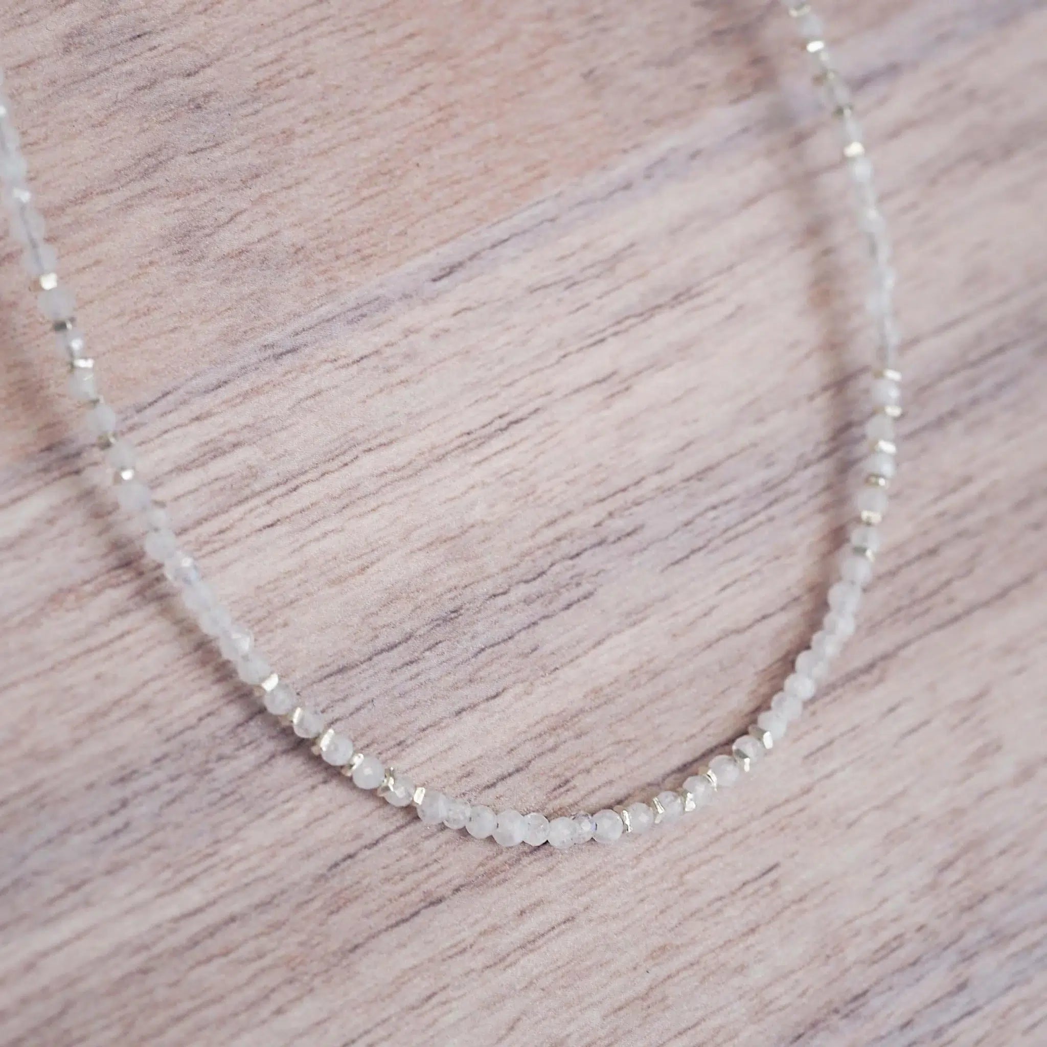 A delicate beaded moonstone necklace made of small, faceted light natural moonstone stones, displayed on a light-colored wooden surface.