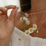 A close-up lifestyle shot of a person holding a delicate silver chain necklace with a central oval pendant featuring a pale blue-gray stone. The person is wearing a white top and holding small white daisies in the background.
