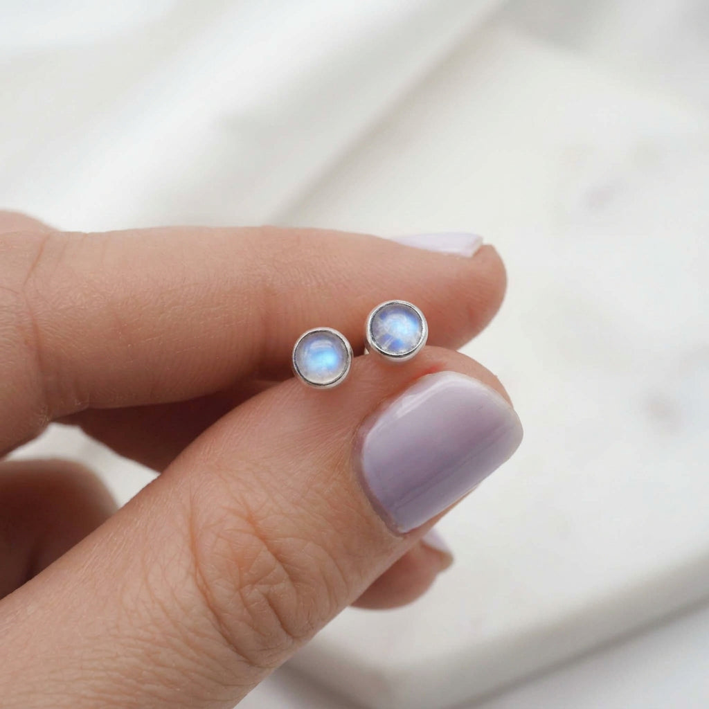 Small moonstone stud earrings in sterling silver held between fingers on a neutral background