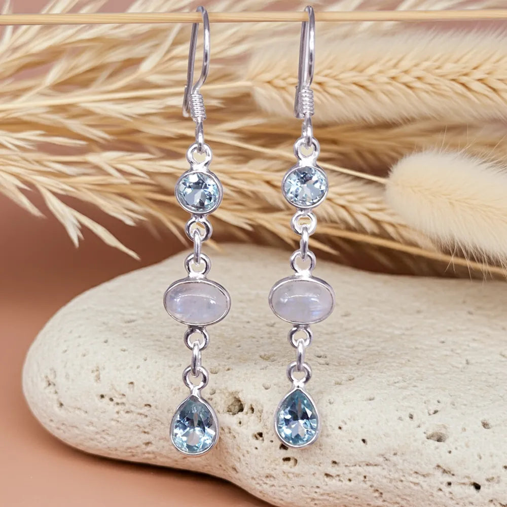 Silver earrings with white topaz and moonstone gemstones on a stone surface with dried grass in the background