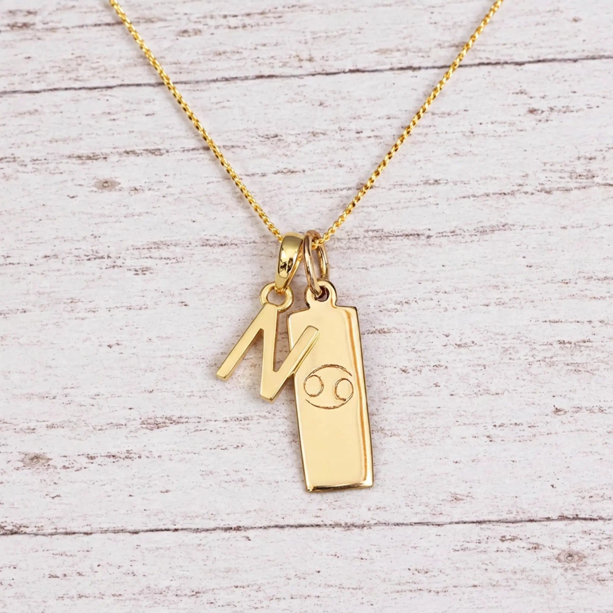 Gold Initial N Pendant and Cancer Zodiac Pendant Necklace on a fine gold chain against a light wooden background