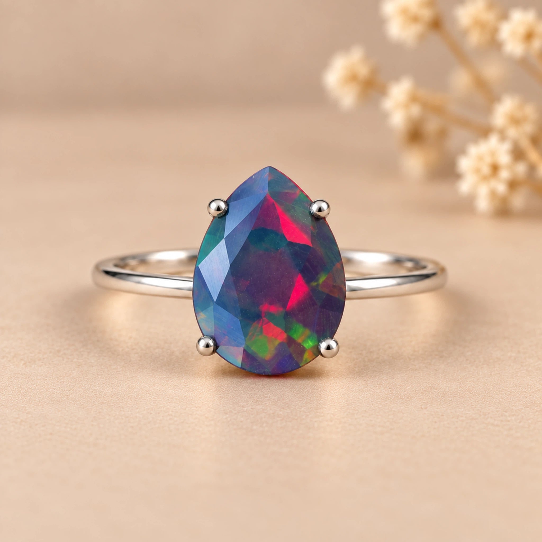 A silver ring featuring a teardrop-shaped iridescent natural australian opal gemstone dyed black that shifts between blue, green, pink, and purple, set against a warm beige background with blurred dried flowers.
