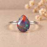 A silver ring featuring a teardrop-shaped opal gemstone with vibrant iridescent colors, set against a warm beige background with blurred dried flowers.