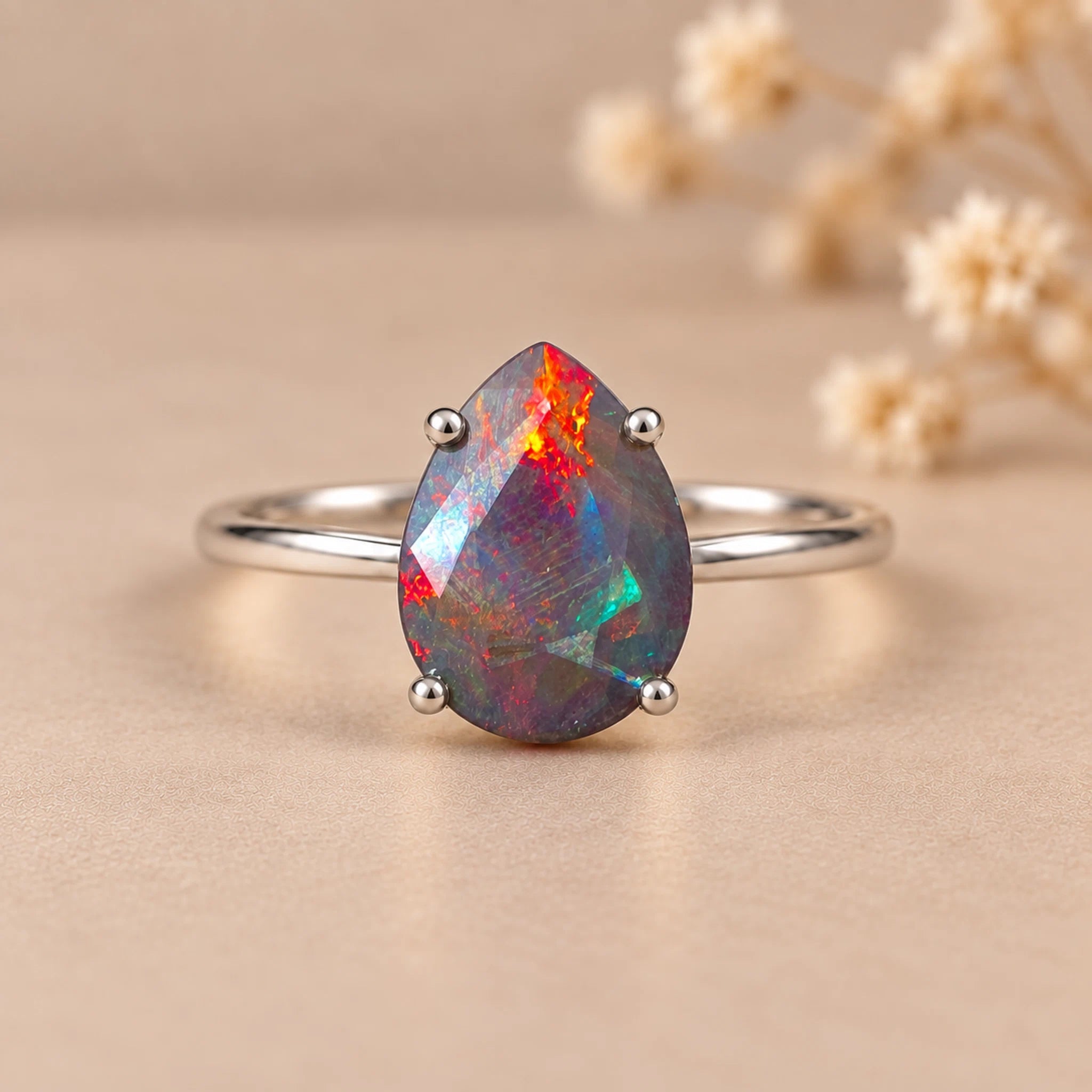 A silver ring featuring a teardrop-shaped opal gemstone with vibrant iridescent colors, set against a warm beige background with blurred dried flowers.