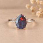 A silver ring featuring a teardrop-shaped faceted opal gemstone with vibrant blue, red, and black iridescent play-of-color, displayed on a warm beige background with soft dried flowers in the background.