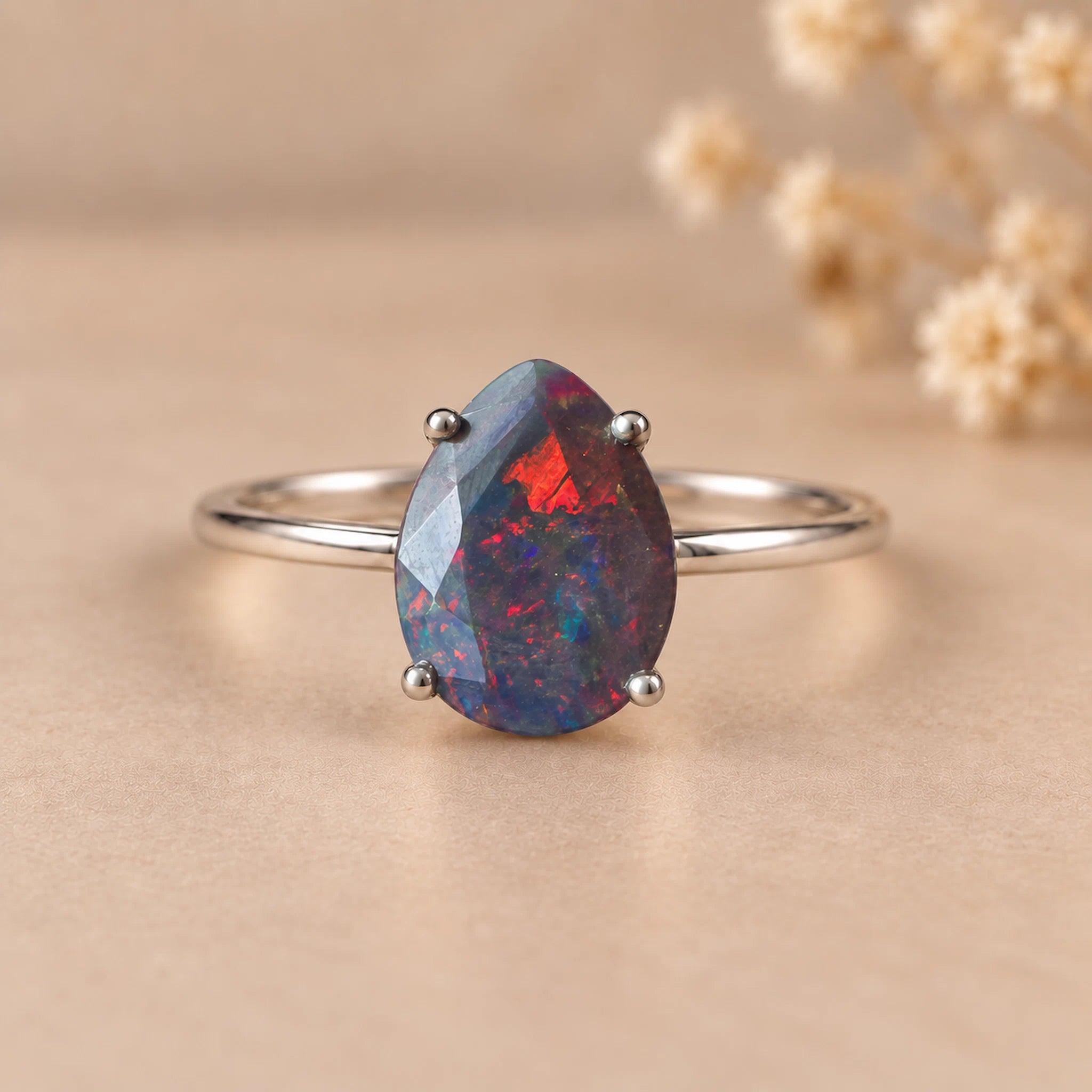 A silver ring featuring a teardrop-shaped faceted opal gemstone with vibrant blue, red, and black iridescent play-of-color, displayed on a warm beige background with soft dried flowers in the background.