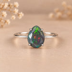 A silver ring featuring a teardrop-shaped iridescent opal gemstone with green, blue, and orange flashes, set on a soft beige background with blurred dried flowers.