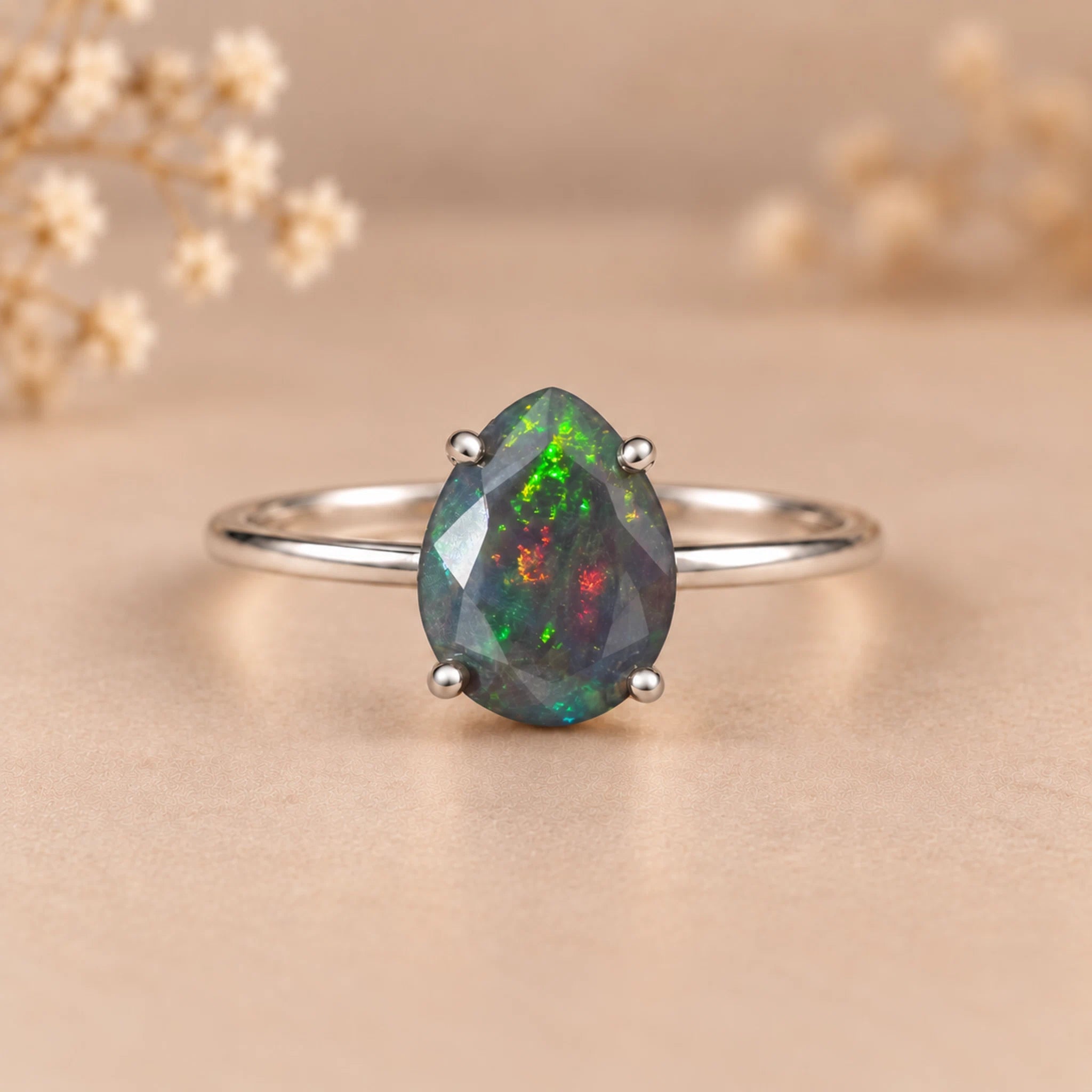 A silver ring featuring a teardrop-shaped iridescent opal gemstone with green, blue, and orange flashes, set on a soft beige background with blurred dried flowers.