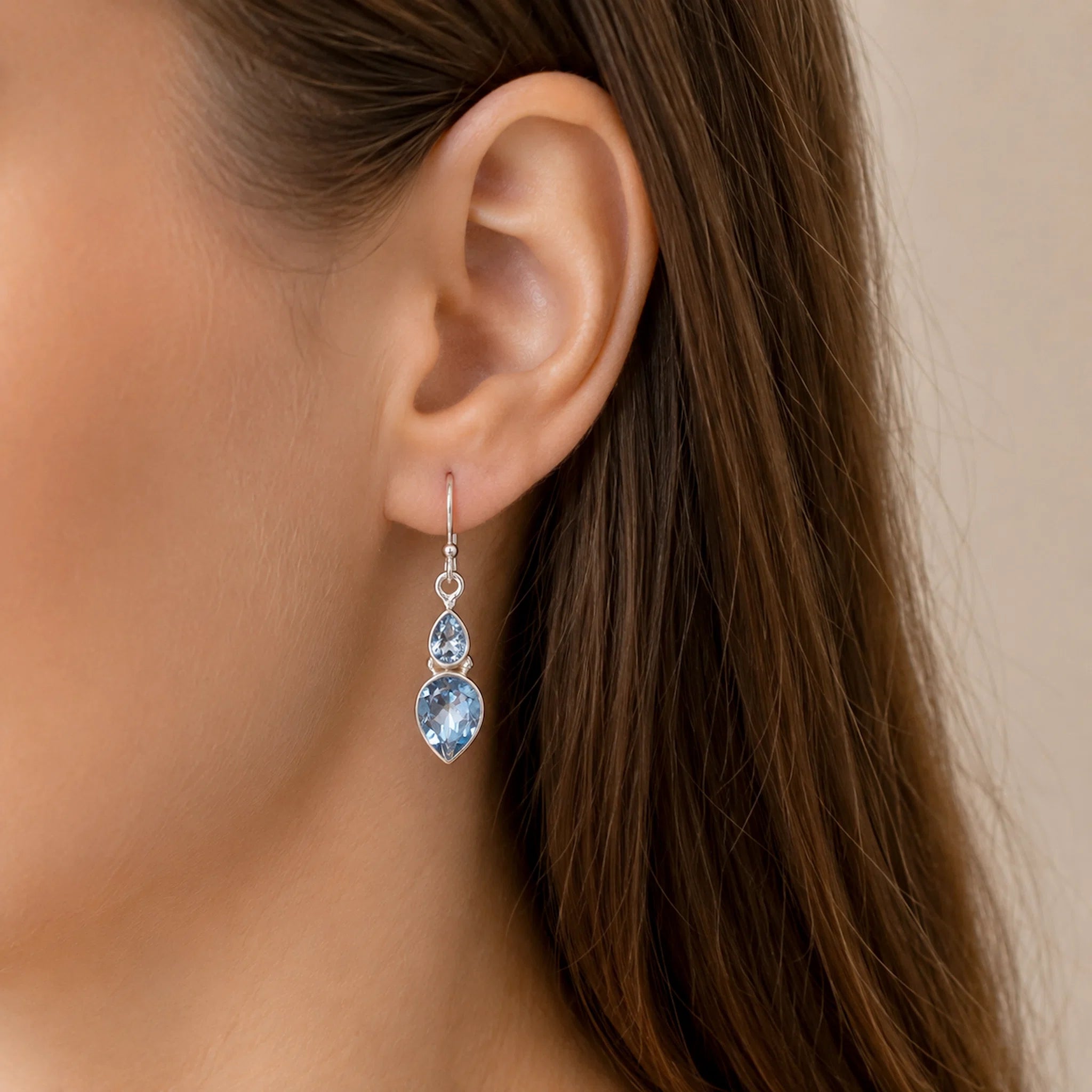 Close-up of an ear wearing a sterling silver french hook earring with two faceted pear-shaped natural blue topaz gemstones with a neutral background