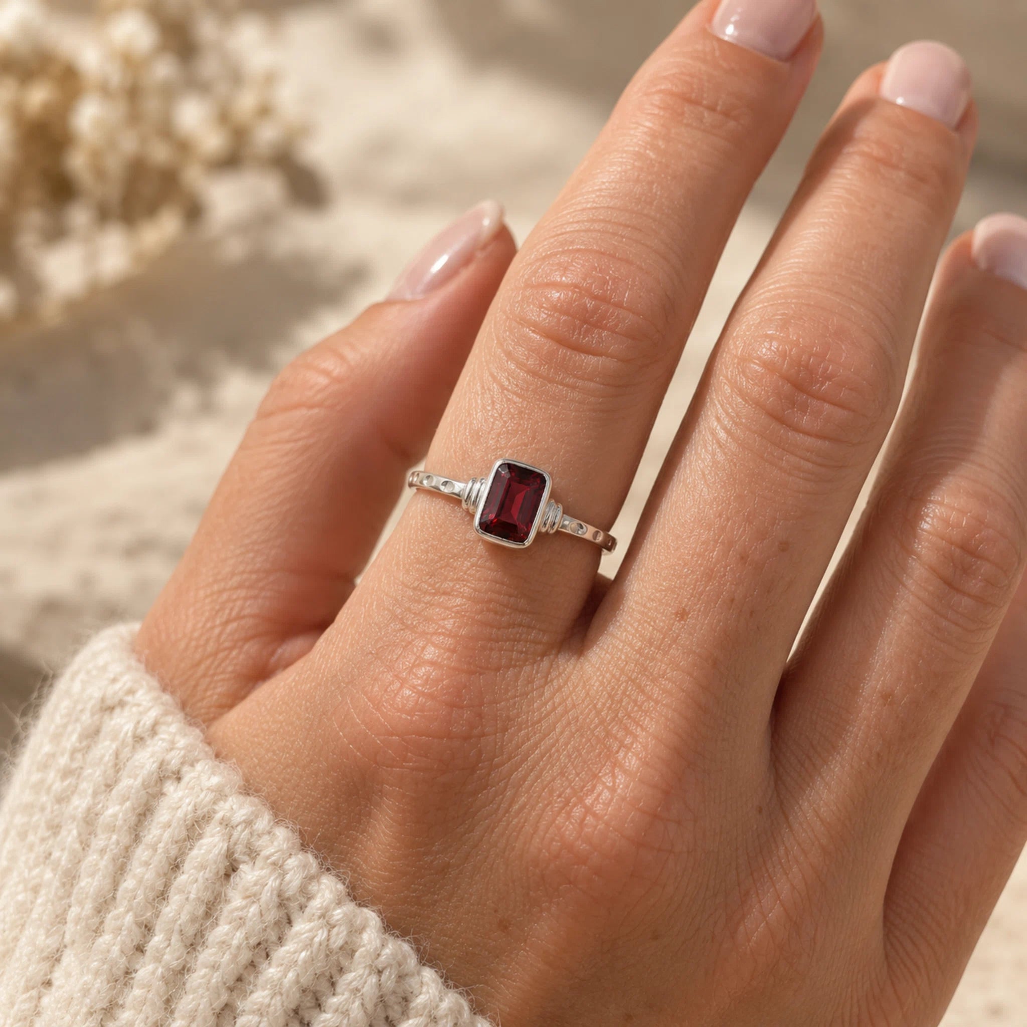 A sterling silver garnet ring with a rectangular emerald-cut bezel-set natural dark red garnet gemstone, worn on a hand with a cream knit sleeve visible, set against a warm, sandy background.