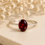 A silver ring with an oval-cut deep red garnet gemstone, displayed on a textured beige surface with dried white flowers in the background.