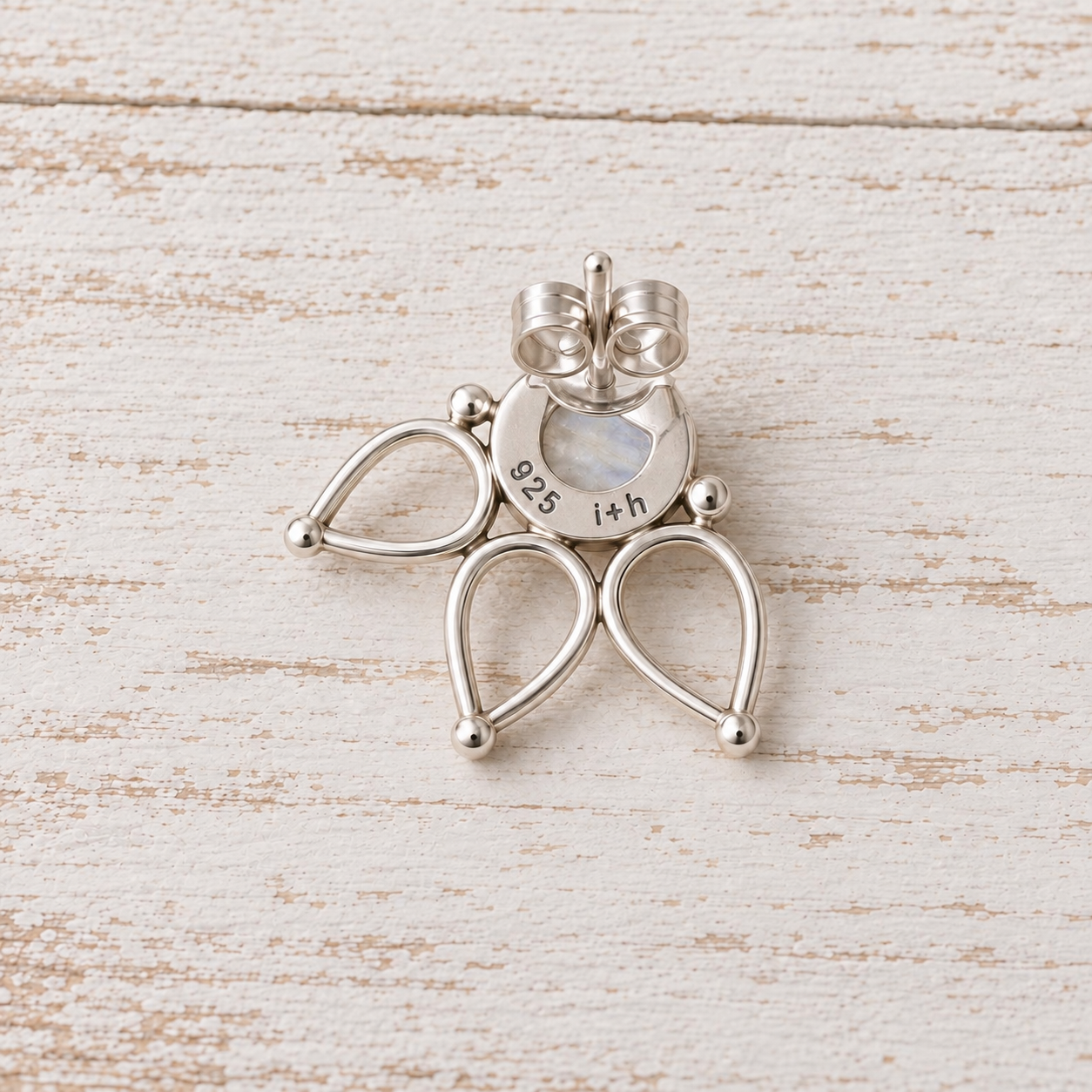A sterling silver butterfly-shaped stud earring with a central round iridescent moonstone, displayed on a distressed white wooden surface.