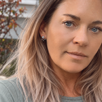 A close-up portrait of a woman with blonde hair wearing a small silver moonstone earring, photographed outdoors with blurred foliage in the background.