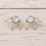 Pair of natural rainbow moonstone lotus flower stud earrings in 925 sterling silver with round cabochon stones, openwork lotus petals, and soft blue flash on a rustic white wood surface.