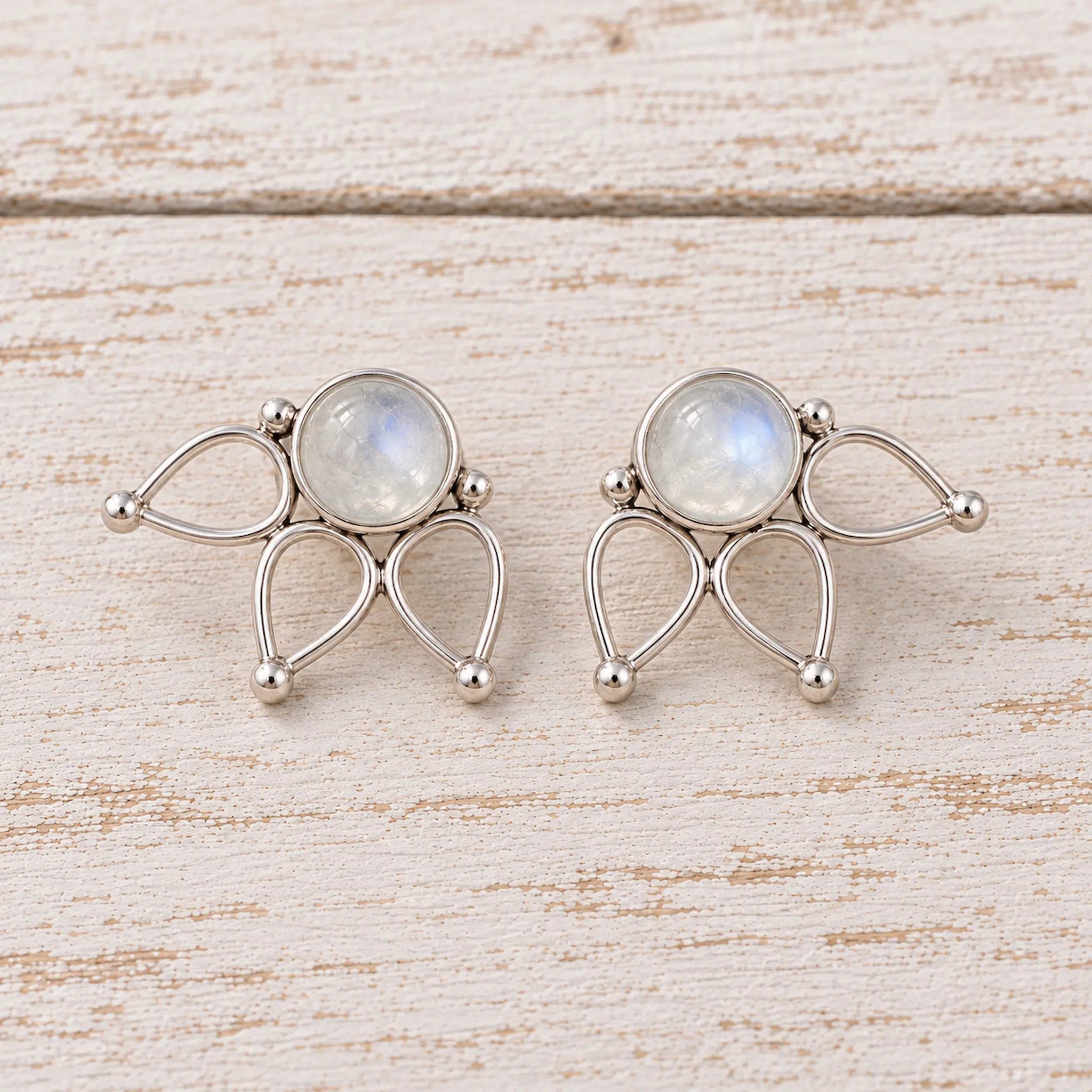 Pair of natural rainbow moonstone lotus flower stud earrings in 925 sterling silver with round cabochon stones, openwork lotus petals, and soft blue flash on a rustic white wood surface.