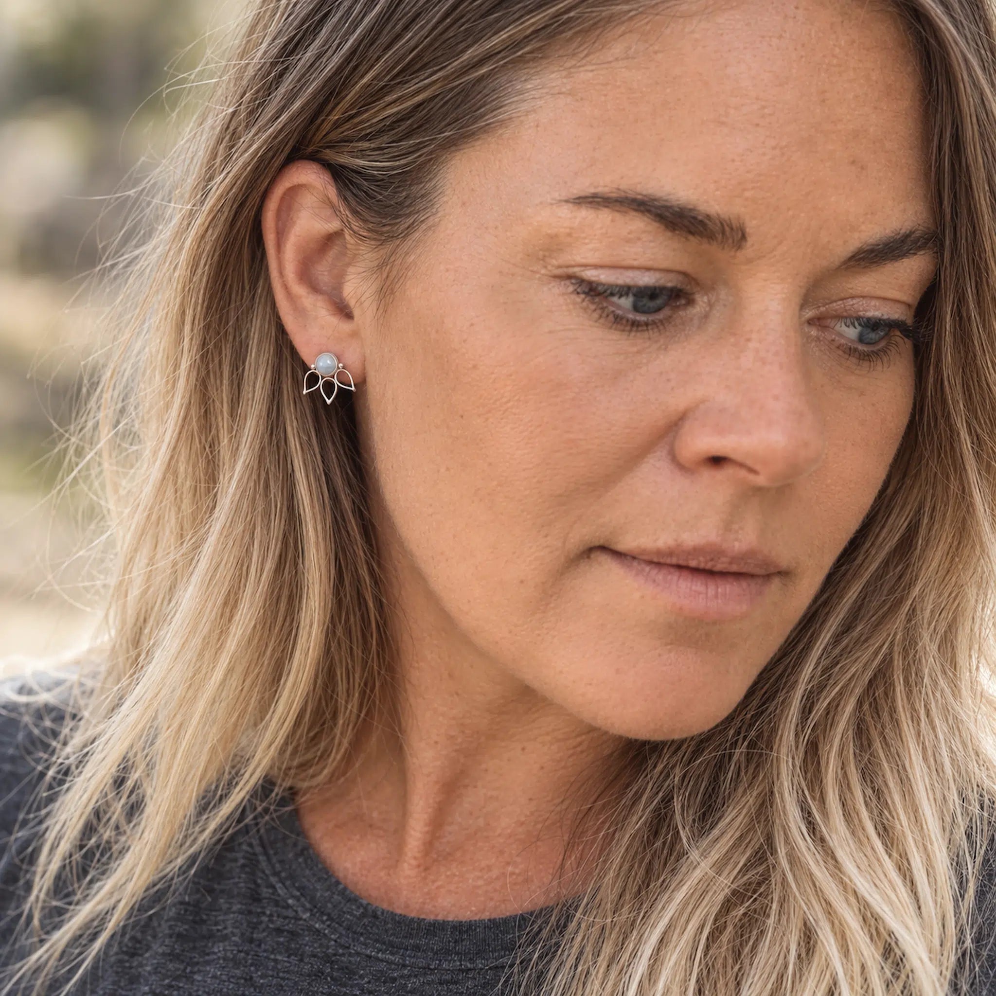 A close-up of a woman with blonde hair wearing a small silver geometric stud earring with a light blue natural moonstone.