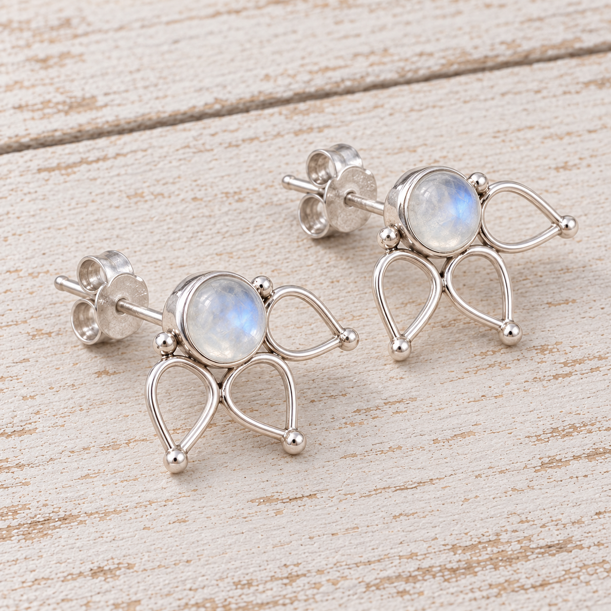A pair of sterling silver stud earrings with teardrop-shaped wirework and round iridescent moonstone centers, displayed on a light wood background.