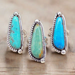 Three silver rings with large teardrop-shaped turquoise and blue-green stone inlays, displayed on a light wood surface.