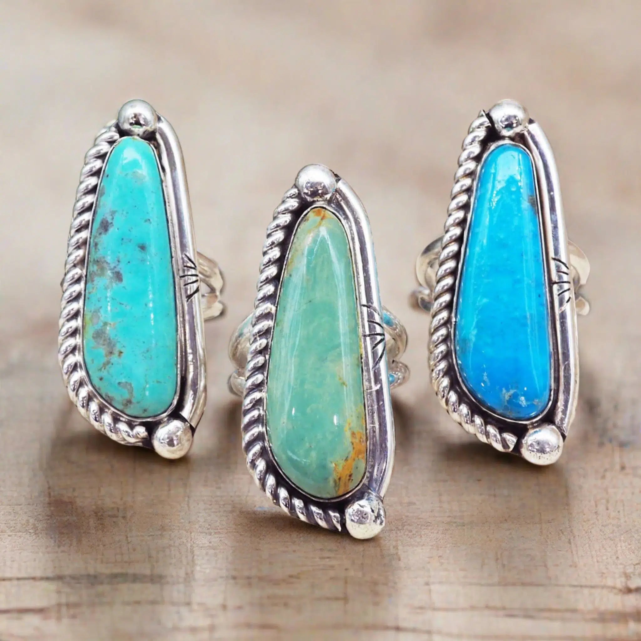 Three silver rings with large teardrop-shaped turquoise and blue-green stone inlays, displayed on a light wood surface.