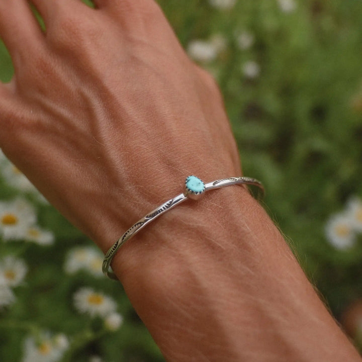 Woman wrist shown over a field of daisies, wearing a silver cuff with a small turquoise stone in the middle.