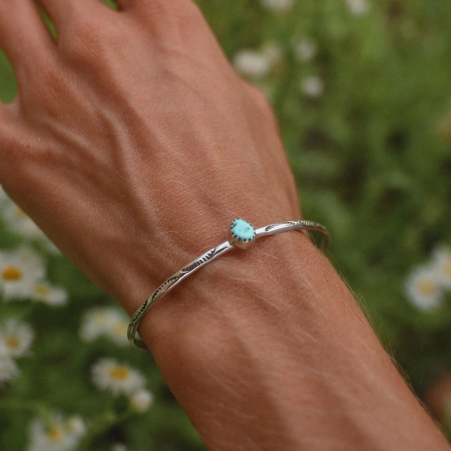 Woman wrist shown over a field of daisies, wearing a silver cuff with a small turquoise stone in the middle.