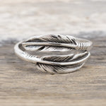 A silver feather ring with detailed feather engravings and a textured band, resting on a rustic wooden surface.