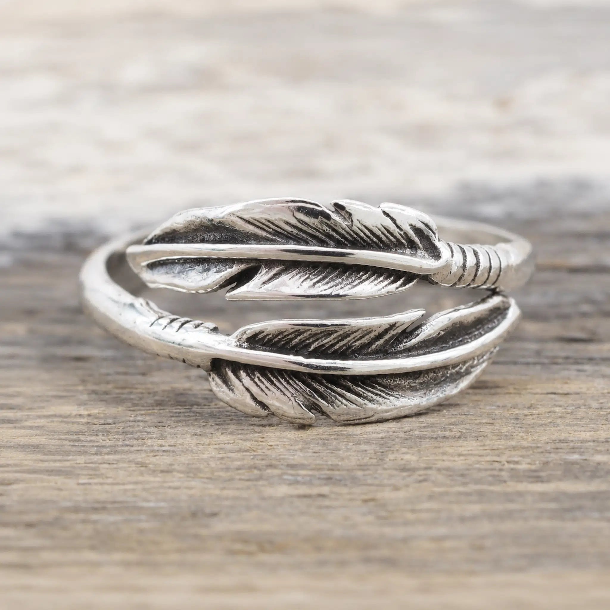 A silver feather ring with detailed feather engravings and a textured band, resting on a rustic wooden surface.