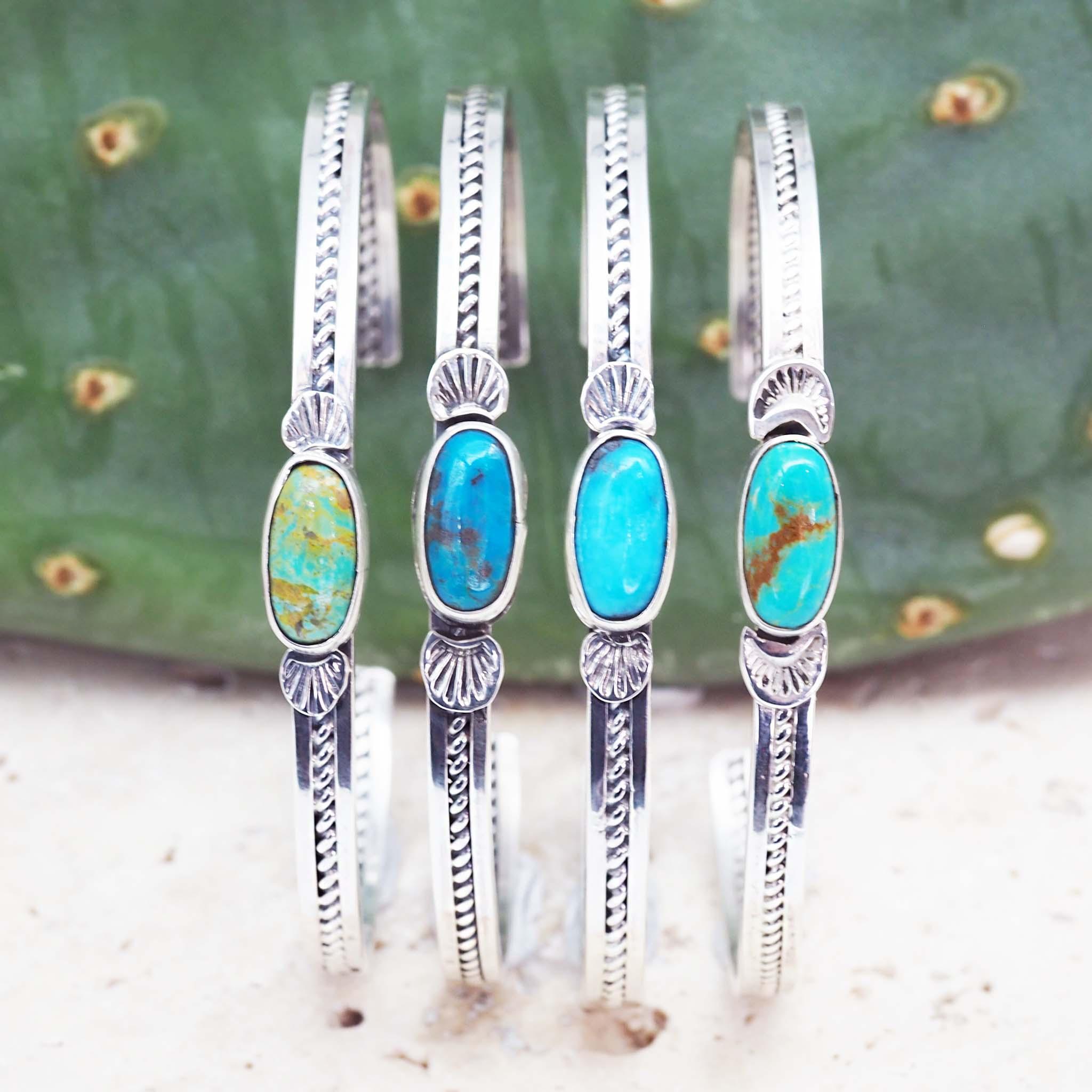 Four handcrafted authentic Navajo turquoise cuff bracelets made of stamped 925 sterling silver with oval natural turquoise gemstones showing colour variations from earthy green to vibrant blue, displayed side-by-side against a green cactus background.