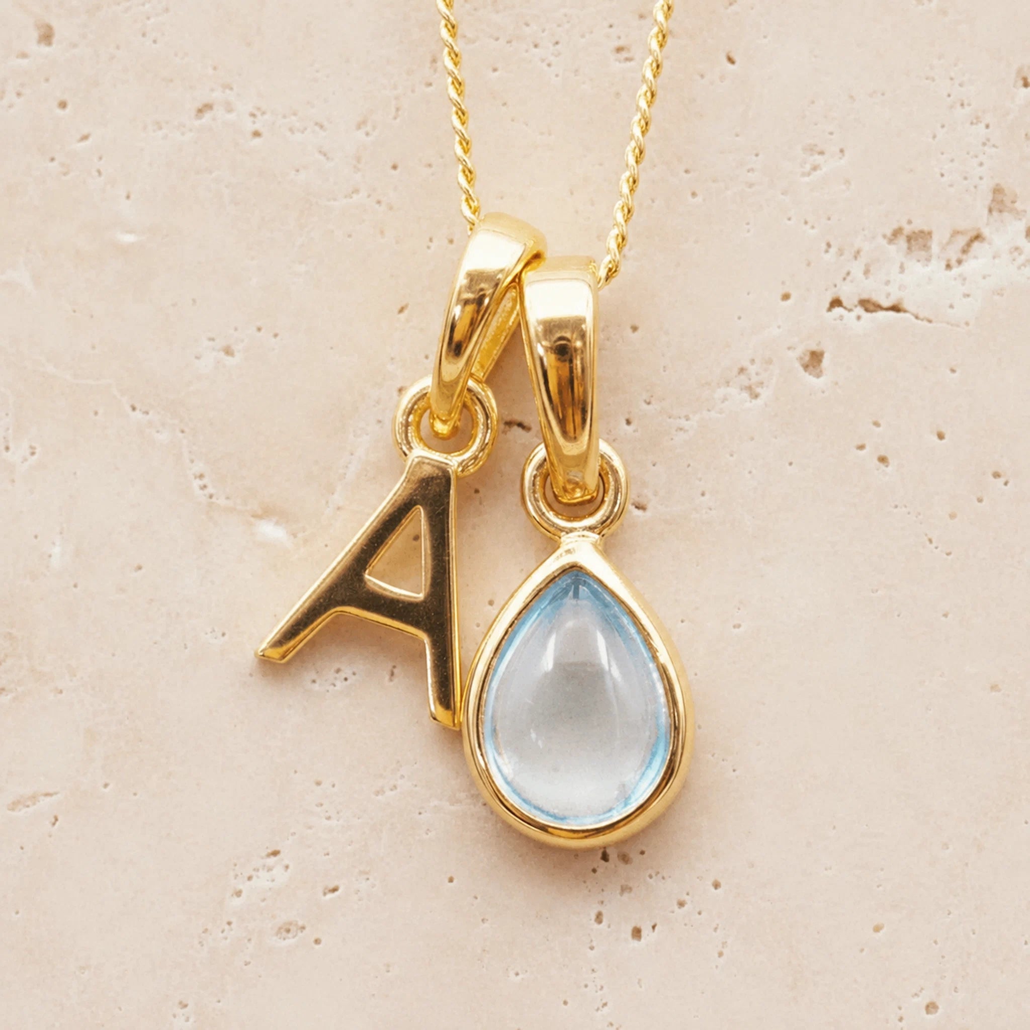 Gold necklace with 'A' pendant and teardrop-shaped november birthstone blue topaz gemstone on a beige background