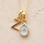 Gold double pendant necklace with a teardrop-shaped november birthstone blue topaz gemstone pendant and a letter Z pendant on a beige background