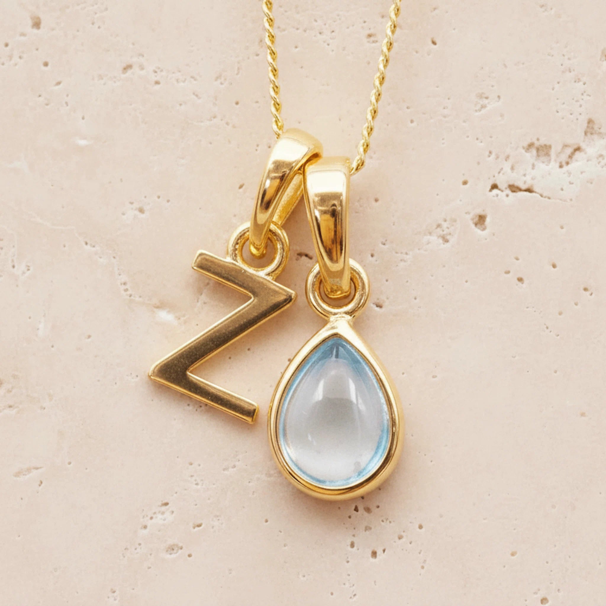 Gold double pendant necklace with a teardrop-shaped november birthstone blue topaz gemstone pendant and a letter Z pendant on a beige background