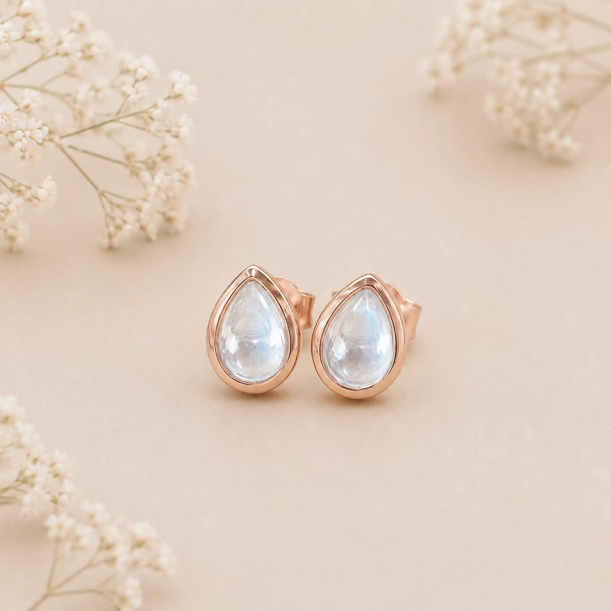 Rose gold november birthstone earrings with light blue topaz gemstones on a beige background with white flowers.