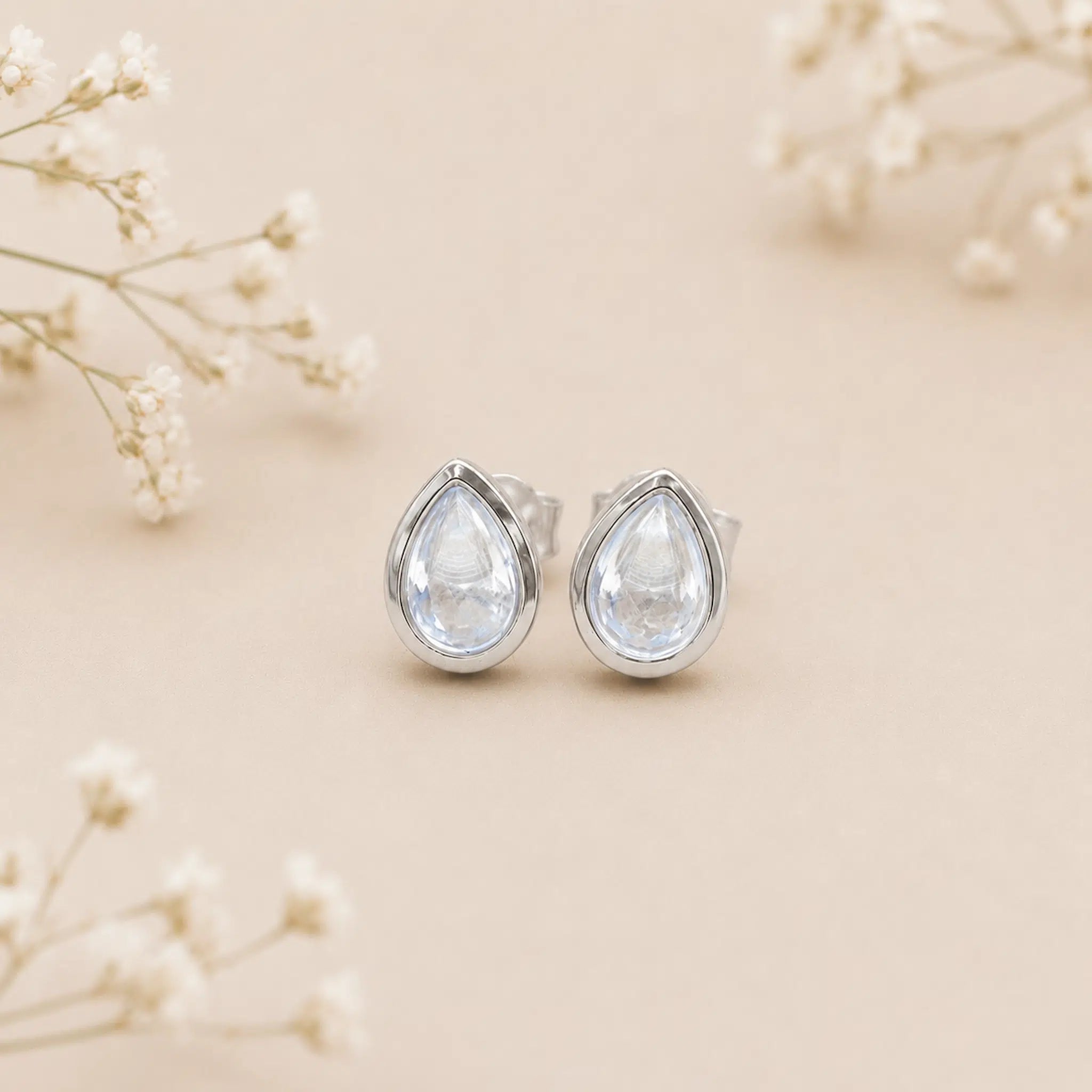 november birthstone earrings with Teardrop-shaped light blue topaz gemstones on a beige background with delicate flowers.