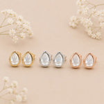 Set of teardrop-shaped earrings in gold, silver, and rose gold on a beige background with white flowers.