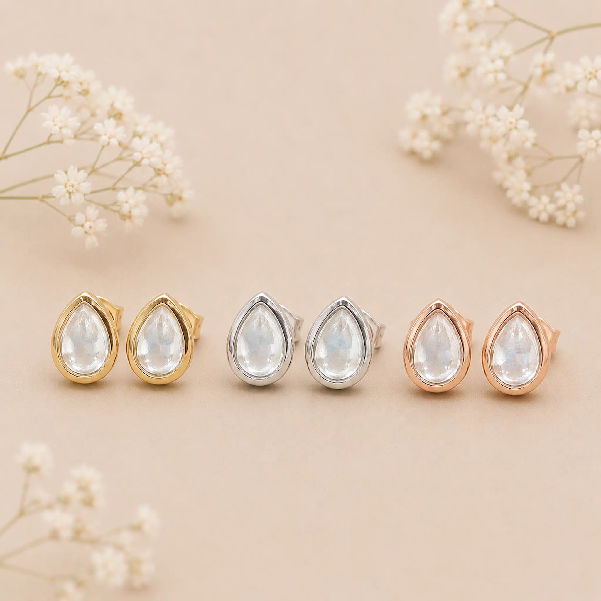 Set of teardrop-shaped earrings in gold, silver, and rose gold on a beige background with white flowers.