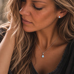close up of a woman wearing a sterling silver November Birthstone Jewellery set featuring a pair of teardrop stud natural blue topaz earrings and matching natural blue topaz pendant necklace 