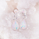 A pair of teardrop-shaped opal dangle earrings with silver settings, displayed on a bed of pale pink crystal clusters.