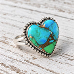 Peacock Dahlia turquoise heart ring with a blue-green stone and gold matrix, angled on whitewashed wood