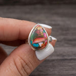 Pink copper turquoise ring held between fingers with white nails, showing the copper matrix, dark wood