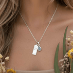 Silver necklace with two charms worn by a person, surrounded by flowers.